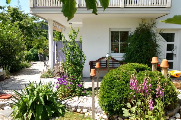 Eine schön gestaltete Gartenfläche zeigt eine Terrasse, darüber einen Balkon, der von einer Säule gestützt wird. Neben der Eingangstür und vor einem weißen Sprossenfenster steht eine Holzbank.