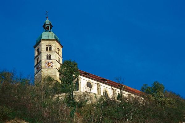 Die Wallfahrtskirche Bogenberg erhebt sich unter blauem, wolkenlosem Himmel. Am Hang darunter dicht wachsende Laubbäume und Sträucher.