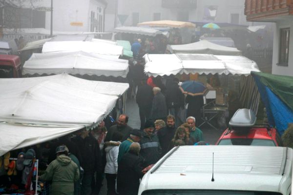 Umgeben von Gebäuden sind mehrere Marktstände unter Pavillons aufgebaut. Dazwischen schlendern Besucherinnen und Besucher. Leichter Nebel liegt über dem Marktplatz.