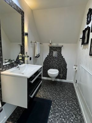 Innenansicht eines Badezimmers mit Dachschräge. Links ein weißes Waschbecken auf einem schwarzen Unterschrank mit großem Wandspiegel. Hinten mittig eine weiße Wandtoilette vor einer dunklen Mosaikfliesenwand. Der Boden ist ebenfalls mit dunklen Mosaikfliesen ausgelegt. Rechts weiße Wände mit schwarzen Deko-Rahmen.