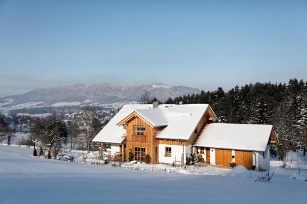 Haus mit Erker und Dachgaube, wunderschön eingebettet in einer Winterlandschaft aus Schnee, Bäumen, Sträuchern und Wald. Im Hintergrund eine verschneite Hügelkette.