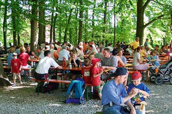 An einem heißen Sommertag kühlen sich die Gäste der Gutsschenke unter Schattenspendenen Bäumen bei einem kühlen Getränk ab. Fast alle Tische des Biergartens sind besetzt.