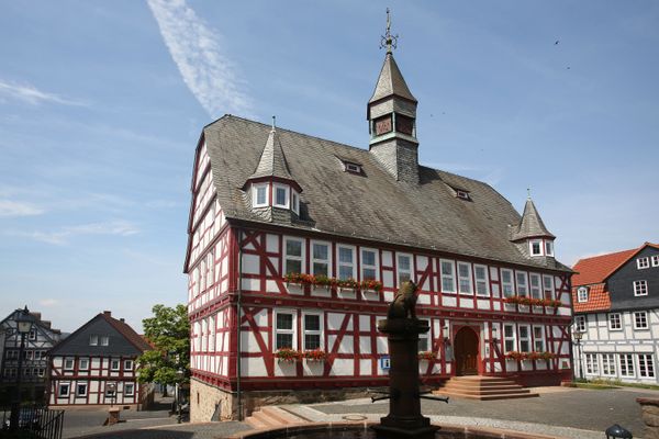 Hinter einem Sandsteinbrunnen erhebt sin ein großer roter Fachwerkbau mit geschiefertem Dach, zwei Spitzgauben und mittigem Turm mit Wetterfahne. Das Fachwerk-Rathaus wurde auf einem Sandsteinsockel gebaut. Neben dem Eingangsportal, das über eine Sandsteintreppe erreichbar ist, hängt ein blaues Tourist-Info-Schild. Vor den weißen Fenstern hängen Blumenkästen mit roten Geranien. Hinter dem Rathaus geht es hangabwäts über eine Treppe zu weiteren, tieferliegenden Fachwerkbauten.