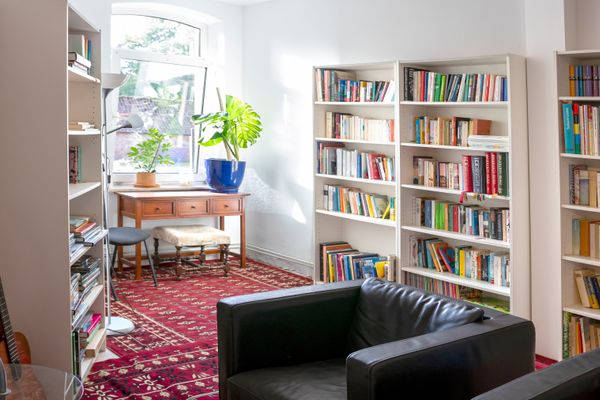 Ein heller Raum mit rotem, gemustertem Teppich, flankiert von hohen, weißen Bücherregalen. Im Vordergrund steht ein schwarzer Ledersessel, im Hintergrund befindet sich ein hölzerner Schreibtisch mit Zimmerpflanzen vor einem Fenster.