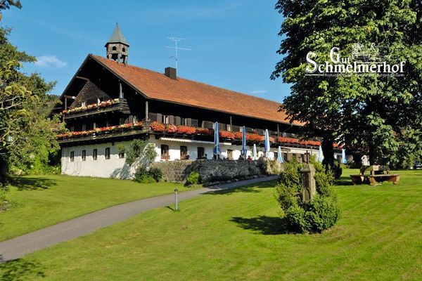 Außenansicht des Hotels Gut Schmelmerhof mit blumengeschmückten Balkonen und Steinterrasse. Das Haus ist umgeben von Bäumen und Sträuchern, die in gepflegten Rasenflächen wachsen.