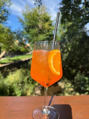 Auf dem Bild ist ein Glas Aperol Spritz mit einer Orangenscheibe auf einem Tisch im Außenbereich stehend zu sehen.