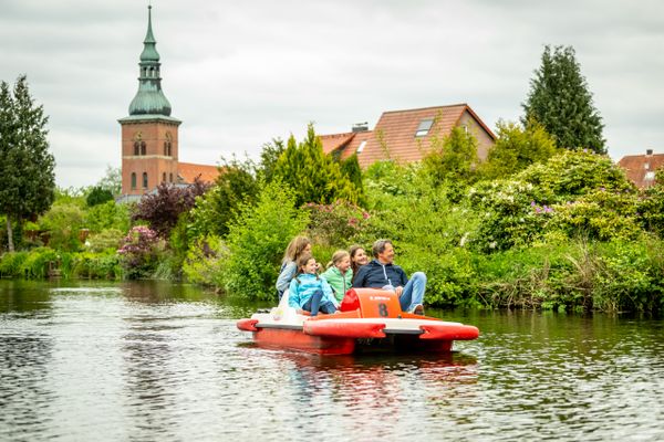 Tretbootfahrer auf See mitbegrüntem Ufer und Kirche im Hintergrund