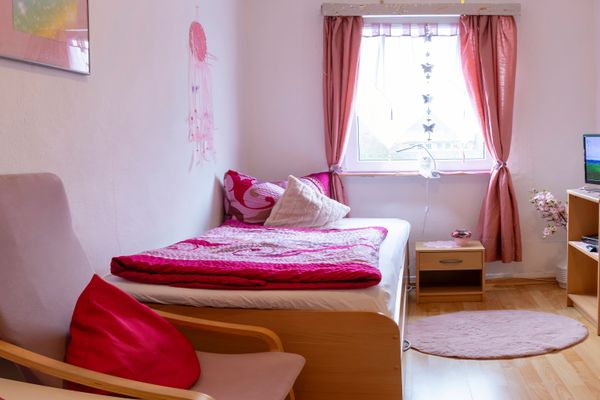 Bett mit rosa Bettwäsche, Fenster mit Vorhängen, Fernseher auf einem Möbelstück und kleiner Teppich