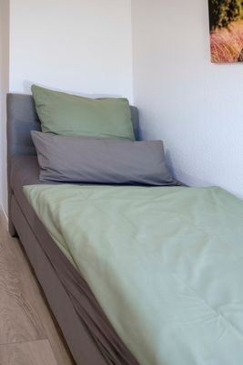 Das Bild zeigt ein Bett in einem modernen Schlafzimmer. Es ist mit einer grauen Matratze, einem hellgrünen Bettlaken und zwei Kissen bezogen – ein hellgrünes und ein graues. Das Bett steht in der Ecke des Raumes, und ein Teil eines Bildes ist an der rechten Wand sichtbar. Der Boden besteht aus hellem Holz.
