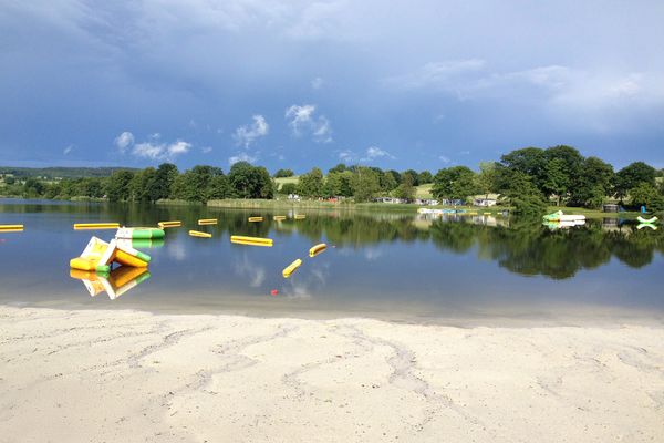 Blick auf den idyllischen Gederner See mit Sandstrand. Auf der gegenüberliegenden Uferseite stehen kleine Gebäude zwischen Baumen und Sträuchern. Ein Wasserspielpark ist auf dem See und die Sonne scheint.