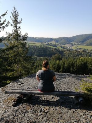 Das Bild zeigt eine Person, die auf einer Bank sitzt. Die Bank steht auf einer Schieferhalde. Von dort hat man einen weiten Blick in die Landschaft.