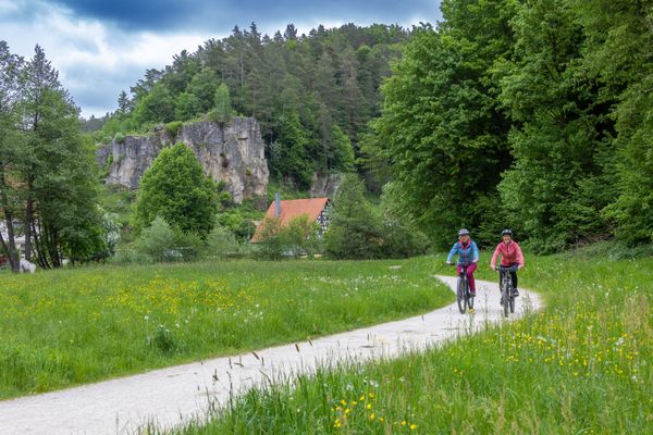 Zwei Frauen die einen Radweg entlang fahren. Im Hintergrund ist ein großer Felsen und ein Fachwerkhaus zu sehen.