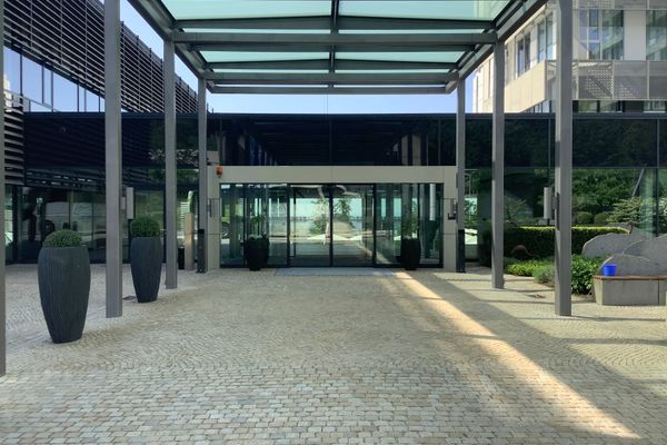 Eingangsbereich eines modernen Gebäudes mit Glasfront, überdachtem Vorplatz und großen Pflanzkübeln.