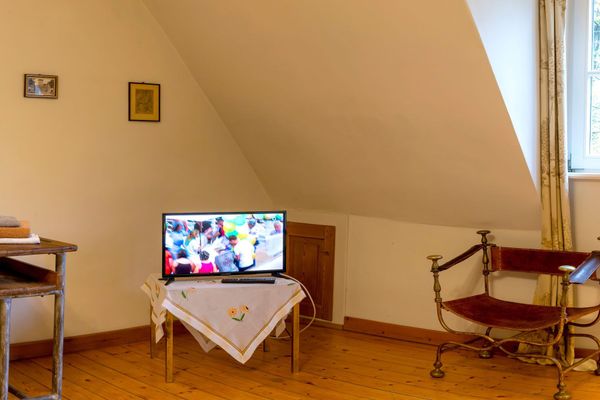 Fernseher auf kleinem Tisch mit Tuch, daneben ein Stuhl und ein Fenster unter einer Dachschräge.