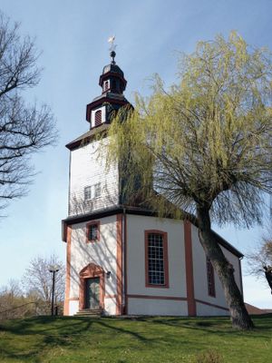 Ein kleines, schlichtes Kirchlein steht zwischen Bäumen auf einer Anhöhe. Der Unterteil mit Kirchenschiff ist weiß verputzt., die Kanten und Fensterlaibungen sind aus rotem Sandstein. Der Kirchturm mit dem achteckigen Haubendachreiter ist dunkelgrau verschiefert. Es ist ein sonniger Tag mit wolkenfreiem Himmel.