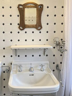Waschbecken mit zwei Armaturen vor einer gefliesten Wand, darüber ein Spiegel und eine schmale Ablage.