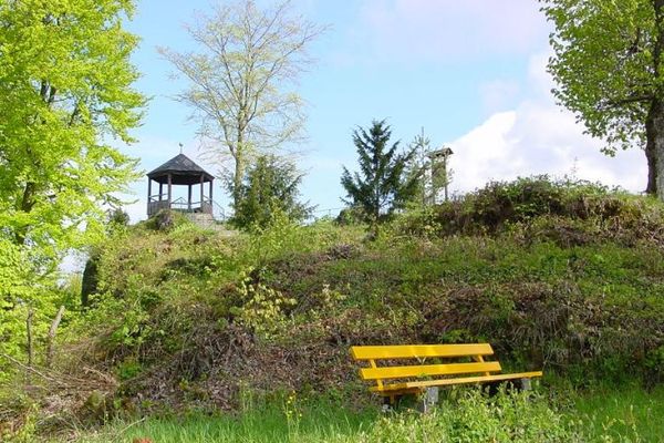Auf einer Anhöhe thront ein hölzerner Aussichtspavillon. Im Vordergrund steht eine gelbe Ruhebank.