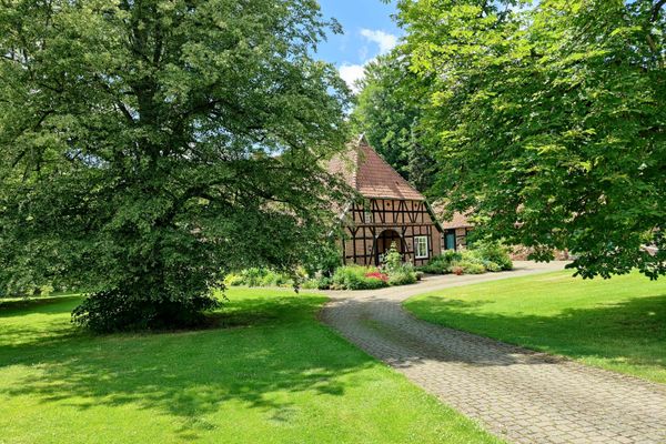 Fachwerkbauernhaus vom Typ niedersächsisches Hallenhaus mit rotem Ziegeldach, umgeben von Rasenflächen, Bäumen und gepflastertem Zufahrtsweg in Böddenstedt unter blauem Himmel.