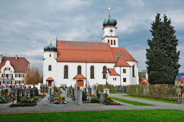 Die Kirche St. Wolfgang in Oberwinkling mit Friedhof. Der Hauptturm ist viereckig mit eckigem Oberbau und Zwiebeldach, gegenüber ein kleinerer Turm mit Zwiebeldach. Dahinter ein schönes, altes Herrschaftshaus mit Sprossenfenstern und Walmdach.