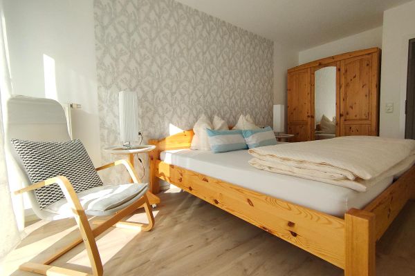 Im Schlafzimmer stehen ein Doppelbett, ein Schrank mit Spiegeltür in der Mitte und ein Stuhl. Neben dem Bett befinden sich zwei runde Tische mit Lampen. Die Möbel sind aus Kiefernholz. Hinter dem Bett ziert eine gemusterte Wand den Raum.