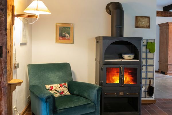 Kaminofen mit Feuer neben grünem Sessel, Lampe an der Wand und Holzbalken im Raum.