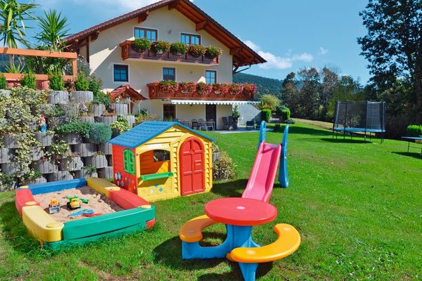 Haus mit Markise über der Terrasse und blumengeschmücktem Balkon im ersten und zweiten Stock. Auf der teils schattigen Liegewiese stehen Trampolin, Rutsche, Spielhaus und Sandkasten. Hinter der Spielecke sind schön bepflanzte Betonringe versetzt übereinander aufgereiht.