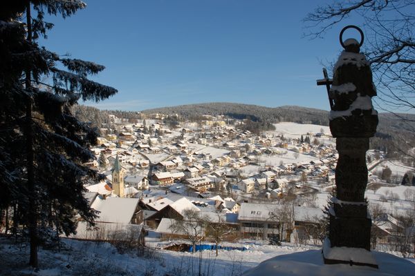 Im Winterwald steht im Vordergrund die Statue des Steinernen Engelmars; im Hintergrund leuchtet das verschneite Sankt Englmar unter der Wintersonne.