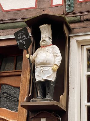 Blick auf ein Detail an einem Fachwerkhaus neben der Eingangstür. Zu sehen ist die Figur eines dicken Kochs, der ein einer Art kleinem, hölzernen Wartehäuschen steht und ein Schild mit der Aufschrift Heute schon gespachtelt in der Hand hält.