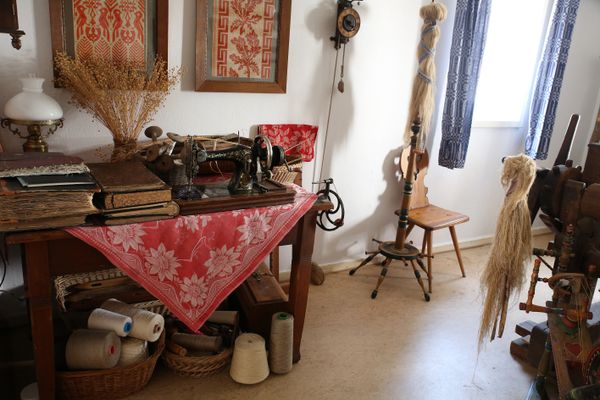 Ein Inneraum des Dorfmuseums zeigt einen Tisch mit Nähmäschine auf einer auffalend roten Tischdecke, darunter allerlei Handarbeitsutensilien. Daneben ist ein Spinnrad zu sehen.