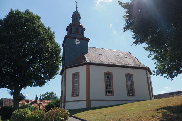 Ausschnitt eines braunen Fachwerkhauses mit Sandsteinsockel. eine einfache, mit Efeu berankte Treppe führt zur schlichten Eingangstür, die oberhalb ein geschnitzter Segensspruch ziert. Es steht geschrieben: Lebe und Liebe, für dieses Haus.