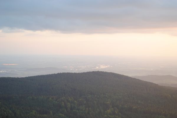 Im Vordergrund eine dicht bewaldete, langgestreckte Hügelkette. Dahinter öffnet sich unter einem bewölkten Himmel der Blick auf eine tieferliegende, weite Landschaft mit einem Ort in der Bildmitte.