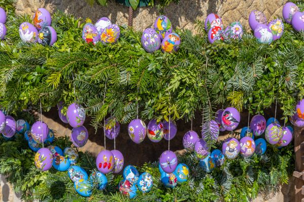 Lilafarbene und blaue Ostereier, die mit Blumen- und Tiermotiven bemalt sind und an Girlanden aus Fichten und Buchsbaumzweigen befestigt sind.