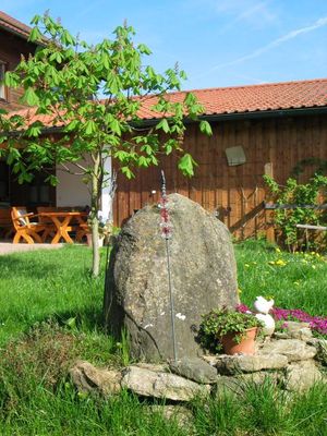 In einer Wiese steht ein mit Bruchsteinen ummauerter und von Gräsern bewachsener Felsen. Im Hintergrund steht eine Sitzgruppe vor einem Holzgebäude.