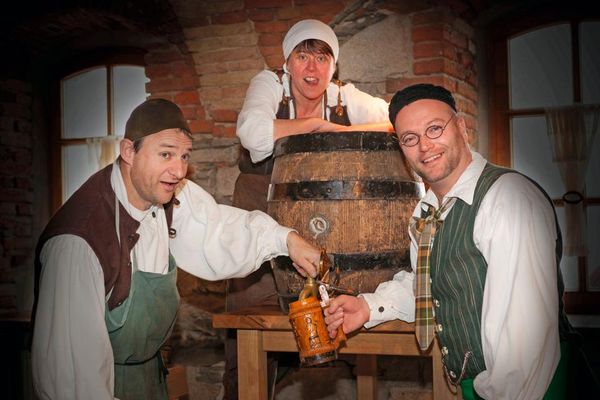 Drei mittelalterlich gekleidete Männer stehen oder lehnen an einem großen Bierfass. Einer hält einen Krug unter den Zapfhahn, ein anderer dreht gerade den Hahn auf.