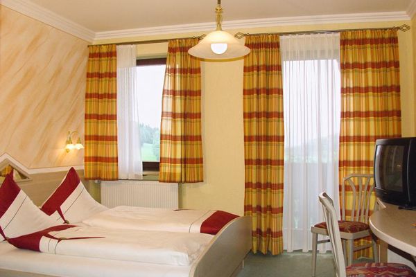 Ein Doppelbett mit hellem Holzrahmen, gegenüber ein Tisch mit zwei Stühlen und ein TV-Gerät. Am Fenster und an der Balkontür hängen Vorhänge mit einem gelb-roten Streifenmuster.