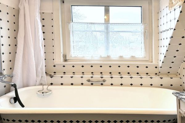 Badewanne unter Fenster mit Vorhang, helle Fliesen und Armatur an der Wand.