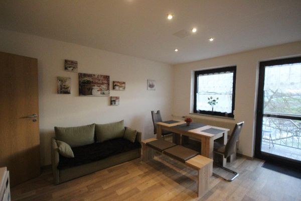 Eine Sitzecke in der Wohnküche mit Holz-Esstisch, zwei Bänken, zwei Stühlen, einem grünen Zweisitzer-Sofa und mehreren Wandbildern. Durch Fenster und Balkontüre fällt Tageslicht.