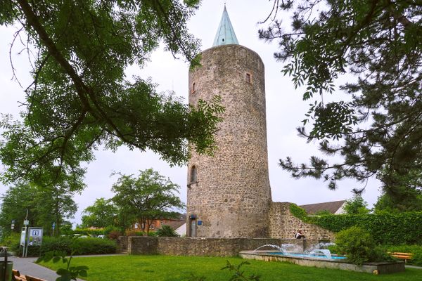 Ein steinerner, runder Turm mit kupfergrüner Dachspitze ist zentrales Bildmotiv. Er ist an niedrige, zum Teil abbrüchige Mauerstücke angebunden und steht auf einer grünen Wiese. Zum Turm führt eine Treppe. Er ist umgeben von Bäumen und es ist herrliches Sommerwetter.