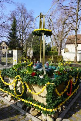 Quadtratischer Brunnentrog, in dessen Zentrum drei bronzene Enten siten, die mit roten Schleifen geschmückt sind. Rund um den Brunnentrog ist eine grüne Girlande aus Nadelhölzern gewunden, geschmückt mit Ketten aus gelben, bemalten Ostereiern. Dazu Frühlingsblumen am Brunnenrand. Über dem Brunnen ein stilisierter Hase als Figur aus Metallstreben, um deren Hals eine Girlande mit gelben Ostereiern.