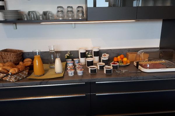 Ein Frühstücksbuffet auf einer dunkelfarbigen Anrichte. Darüber befinden sich ordentlich aufgereiht Teller und Gläser.