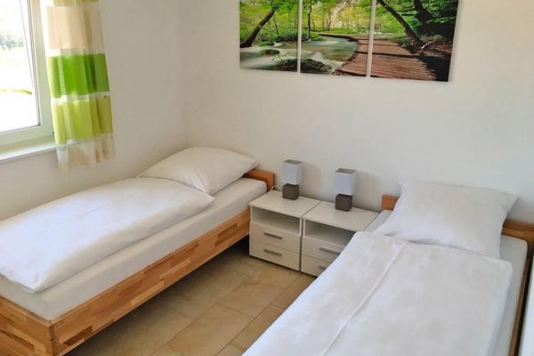 Ein Schlafzimmer mit zwei auseinander stehenden Betten auf Holzgestellen, dazwischen stehen zwei weiße Nachtkästchen. Über den Betten hängt ein dreiteiliges Bild, das einen Laubwald mit einem Bach und einem breiten Holzsteg zeigt.