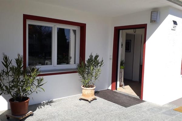 Vor einem Fenster und der offenen Haustür stehen Grünpflanzen. Die Türen und Fenster sind rot umrahmt.