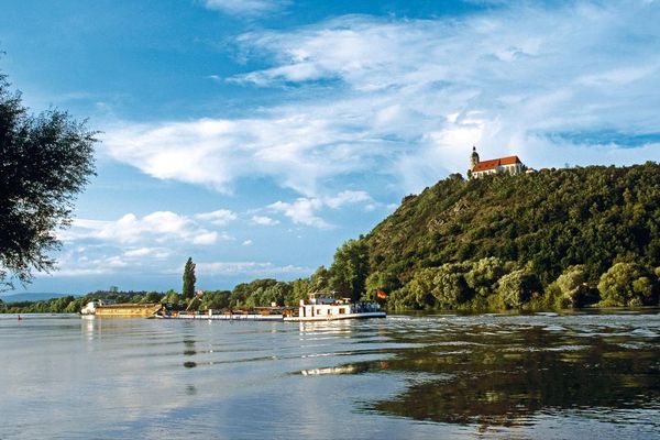 Ein Lastschiff fährt auf der Donau, darüber erhebt sich der Bogenberg mit der Wallfahrtskirche. Der Laubwald spiegelt sich im Wasser.