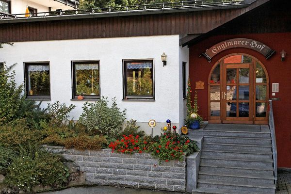 Eine Steintreppe führt zur runden Eingangstür mit Sprossenfenstern. Über der Tür steht „Englmarer Hof“. Vor dem Haus befindet sich eine Steinmauer und ein Beet mit Blumen und Sträuchern.