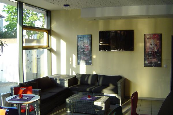 Cafeteria mit Sitzgruppe aus grauem Rattan-Sofa und Hockern, zwei kleinen Tischen mit roten Stühlen, großem Fenster und zwei an der Wand hängenden Fernsehern