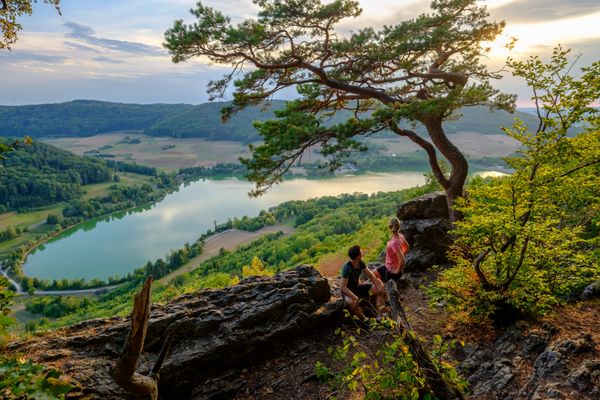 Ein Mann und eine Frau stehen am Aussichtspunkt Hohler Fels auf der Houbirg. Das Bild zeigt einen weiten Blick auf den Happurger Stausee und die dahinter liegenden Hügel. Der Vordergrund zeigt die zwei Personen sowie kleinere Felsen und einen Baum.