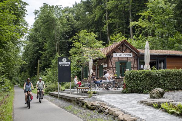 Radfahrer fahren auf dem geteerten Vulkanradweg an der Zentralstation in Lauterbach-Frischborn vorbei, die mitten in einem Waldstück liegt. Die Zentralstation ist der ehemalige Bahnhof und wird heute gastronomisch mit einem Biergarten genutzt.