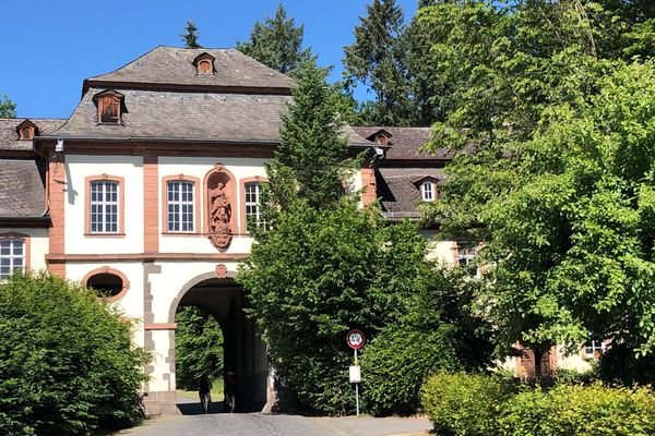 Der Ausschnitt eines Barockbaus mit Durchfahrtsbogen. Der weiße Bau mit Sandsteinen an den Kanten und zur Verzierung hat ein Schieferwalmdach. Über dem Torbogen ist ein Sandsteinbogen mit Ornamenten auffalendes Schmuckelement.