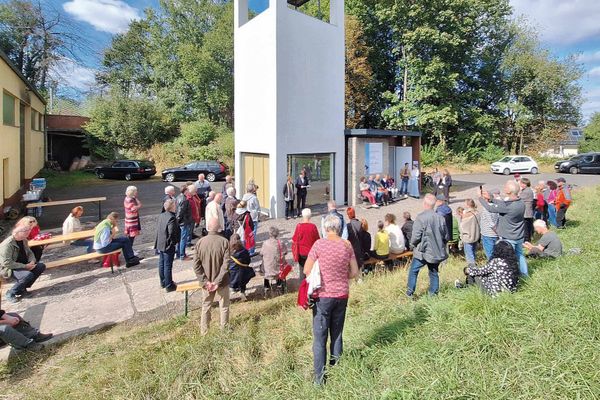Rund um einen markanten, weißen Turm mit quadratischer Grundfläche und bodentiefen Fenstern an jeder Seite im Erdgeschoss stehen vielen Menschen. Manche sitzen auf Brauereigarnituren oder auf einer Bank an einer Betonwand rechts neben dem Turm. Zwei Personen scheinen unmittelbar vor dem Turm einen Vortrag zu halten, die anderen blicken in ihre Richtung. Im Vordergrund ist eine Wiese, hinter dem Turm stehen ein paar Autos vor  ein paar Bäuemen. Am linken Bildrand ist eine Wand eines gelben Gebäudes zu sehen. Es ist schönes Wetter.