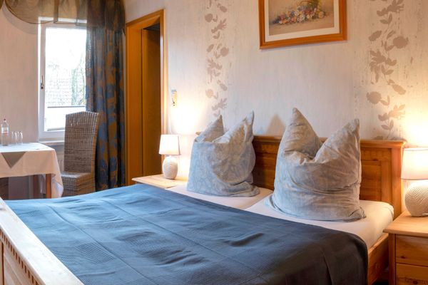Ein Schlafzimmer mit einem Doppelbett aus hellem Holz, bedeckt mit einer blauen Tagesdecke und zwei aufgestellten Kissen. Links steht ein Tisch mit zwei Stühlen vor einem Fenster mit blauen Vorhängen. Auf den hölzernen Nachttischen rechts und links des Bettes leuchten zwei Lampen.